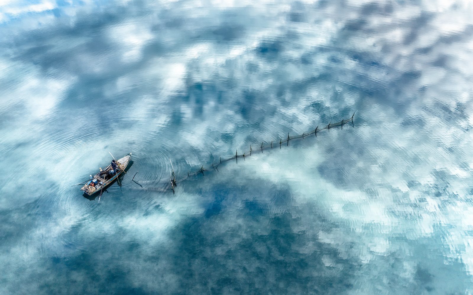 Veduta dall’alto di una barca nella laguna, con una fila di pali e nuvole riflesse nell’acqua