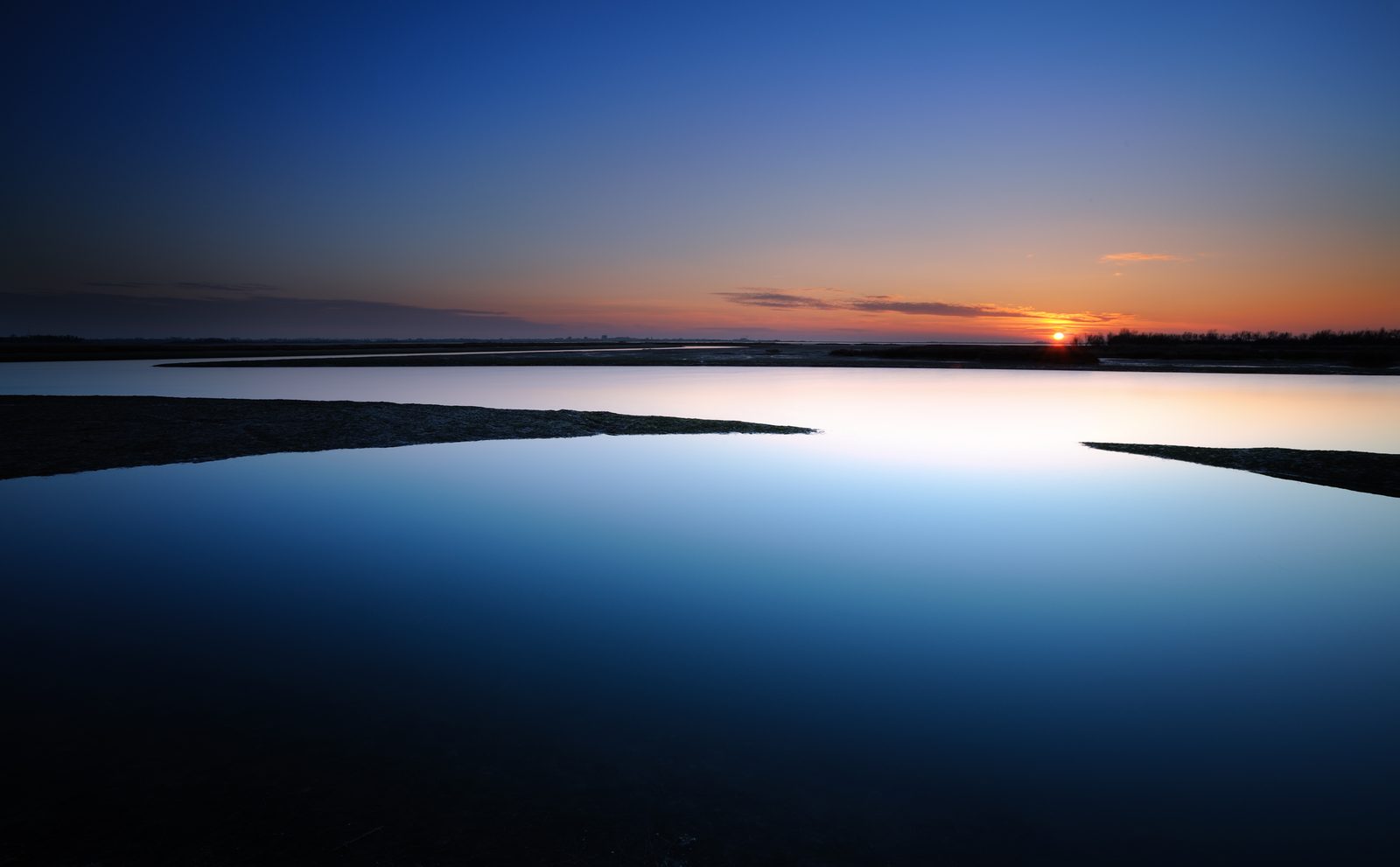 Il sole basso sulla laguna, con barene sottili e acqua blu profondo